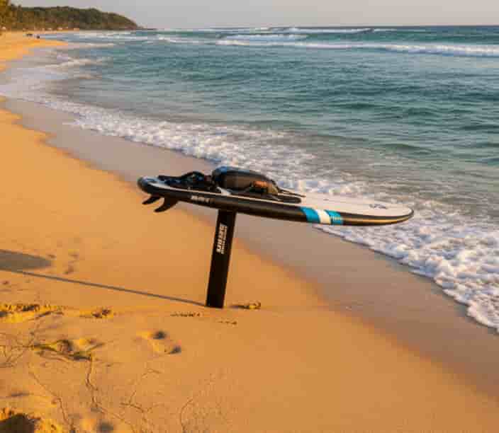 E-foil surfboard resting on sandy beach with ocean waves in the background