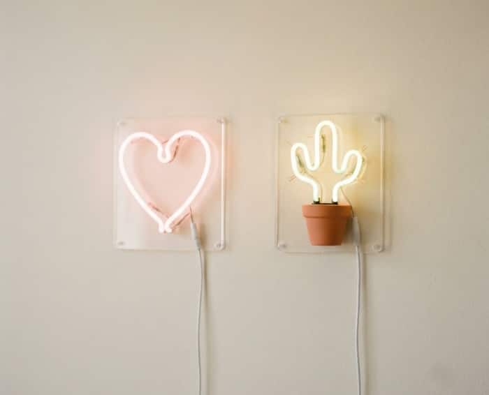 Two handmade neon signs, a pale pink heart and a soft green cactus, hanging on a plain light cream wall