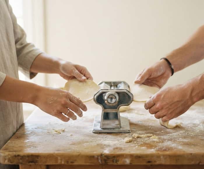Two pairs of hands carefully feeding fresh dough through a vintage hand-crank pasta machine on a flour-dusted counter