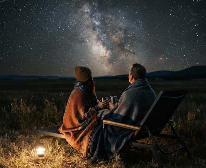 A couple wrapped in blankets sitting on lawn chairs, stargazing under the Milky Way on a clear night.