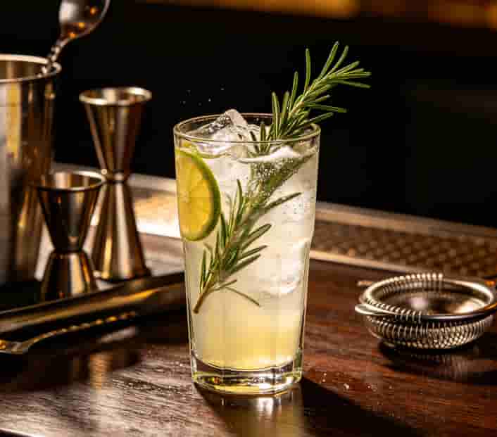 Zero-proof cocktail in a glass with rosemary, citrus, and ice, next to bar tools on a counter
