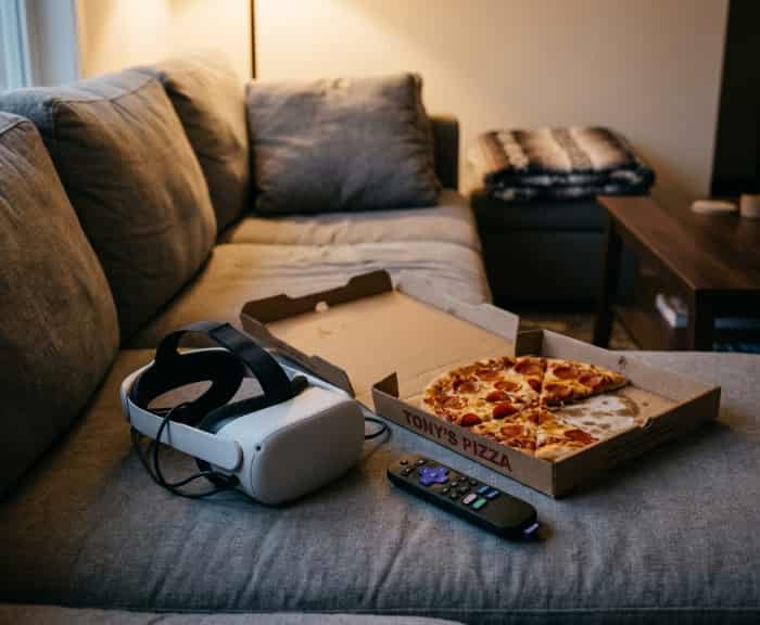A VR headset on a grey couch cushion next to an open pizza box and a remote control under warm evening light