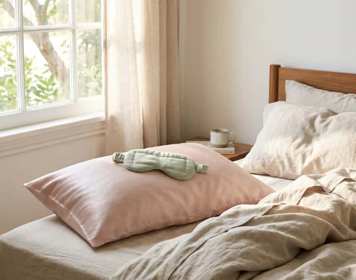 A pastel pink silk pillowcase and green weighted eye mask on a neutral linen bed by a sunlit bedroom window
