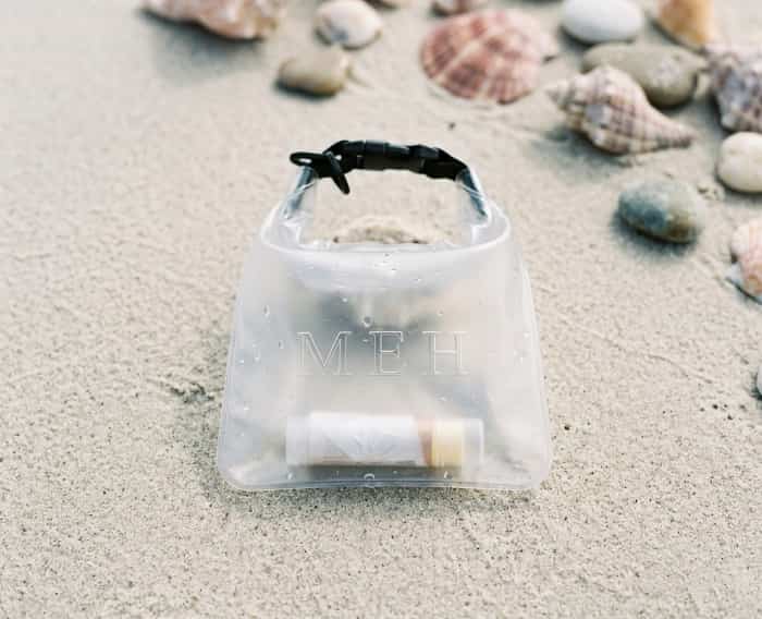A clear waterproof pouch with embossed initials on a sandy beach surrounded by seashells