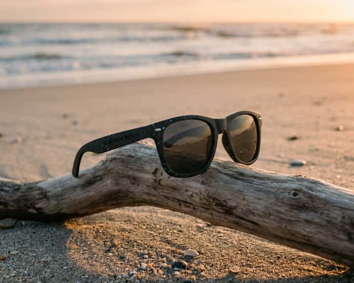 Matte black sunglasses made from recycled ocean plastic resting on driftwood at a beach sunset