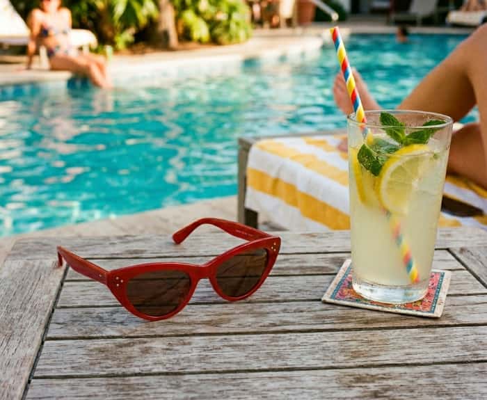 Retro cherry red cat eye sunglasses on wooden table next to glass of lemonade with poolside background