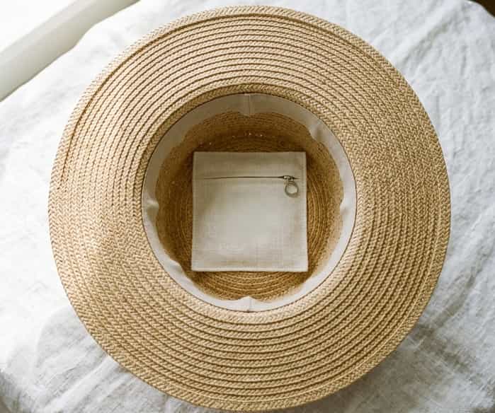 An upside-down wide-brim straw sun hat on a white linen cloth, revealing a hidden zippered fabric pocket inside